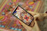 The artist is holding a phone and looking at a colourful sculpture made of candy. On the phone, you can see a digital version of the artwork.