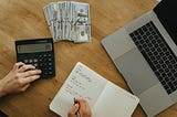 Here we have a picture of a person calculating all of the money that they made writing, just like I did for this article. There are disembodied hands over a calculator and notepad. A stack of money and laptop is nearby on a wooden table.