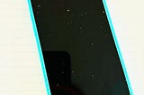 This is a photo of a cell phone (Apple iPhone) with a white background, a turquoise phone case that borders a black screen that shows a bit of dust on it.
