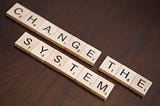 Scrabble tiles spelling out “change the system” sit on a wooden table.