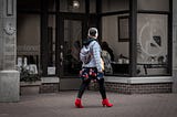 Photograph of a young woman walking on a sidewalk past a store. She is elegantly dressed and wears bright red shoes