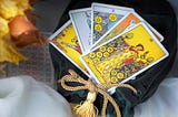 A fanned spread of 5 Tarot cards sitting on a green velvet bag tied with a golden cord. The bag sits on a table decorated in orange flowers and a white linen cloth at the bottom of the image.