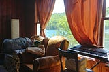 A living room window looks out to a lake surrounded by trees