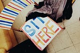 Bird’s eye view of wooden and metal stack chair painted in white, red, orange and blue, with text on seat: “SIT HERE”.