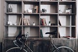 Photo of a gray bookshelf with books sparsely spaced and a road bicycle leaning in front of it. Neutral toned and relaxing to look at.