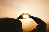 Person holding fingers up in a heart shape with sun shining through.
