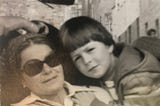 A woman wearing sunglasses sitting in a car, a young child leaning in close, with his hand on her shoulder