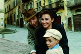 A man with a hat on his bicycle. His beautiful wife and his young son sit in front of him. They are all happy carrying out with their daily lives.