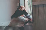 A woman leans against a windowsill in quiet contemplation, resting her head on her hand. Soft light filters through an open wooden window, creating a calm, introspective atmosphere.