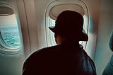An elderly woman wearing a hat and looking out a window of a plane descending for landing.