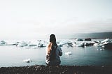 A girl sitting with her back to us on a black beach in front of water filled with iceberg pieces. A moody setting.
