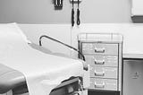 BW photo of doctors office, empty exam table and storage drawers.