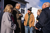Gov. Jay Inslee and Trudi are standing outside bundled up in warm jackets, chatting with a man in a stocking cap at a Longview tiny home village. The sun is shining while they talk.