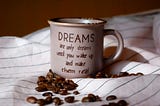 A white coffee cup sits on striped sheets and has a quote that reads, “Dreams are only dreams until you wake up and make them real”.