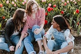 ID: Three women — two light skinned, the last one dark skinned — sit in the ground outside. There is a field of pink and red flowers behind them.