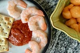 shrimp cocktail with saltines and cocktail sauce on a plate on a gray marble counter next to a green bowl of canteloupe balls