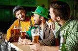 A group of three young men sit at a bar with pints of beer at the ready. They are smiling and obviously having a good time. Two are wearing hats; one of the hats is bright Irish green and sports a shamrock.