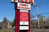 The photo shows a free-standing sign in front of Town Spa, a restaurant in Stoughton, Massachusetts. Town Spa is at the top in white letters, below it is pizza spelled out in red. The bottom part of the tall sign lists other food items and their telephone number.