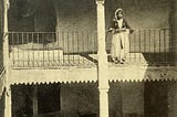 1912 black and white postcard of a two storey moorish house with proches. A woman stands on the second floor balcony. She is wearing a traditional costume with a short jacket and flowing skirt