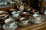 Photograph of multiple white and painted china teacups sitting on china saucers on a brown wooden sideboard, with a gold teapot in the background.