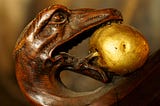 Finely detailed wood carving of a snake with human eyes, holding in its mouth, as if in presentation, a gold-colored fruit