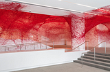 Standard white walls and concrete floor of a modern museum lobby. Stairs are to the right and there’s a central column. Along the walls, ceiling, and even the column is red yarn, like a massive, messy spiderweb.