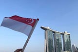 a hand held falg of Singapore waving against the backdrop of the Marina Bay Sands Hotel