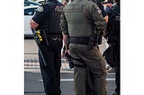 Two police officers, center frame, stand facing away from the camera. One has his hand on gun.