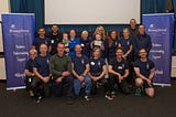 16 people in a group shot (two rows, front row are kneeling) made up of MDP fitness instructors, Ali, and Carl and Jen with their babies, either side is a BleepKind roll up banner and there is a projector screen in the background