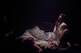 Still from “A Banquet” featuring Betsy played by Jessica Alexander and Holly played Sienna Guillory. Both are in a dark room with a single light overhead. Holly is praying while Betsy is on her back screaming.