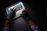 Image of a laptop with two hands typing on it using burglar-like black hand gloves.