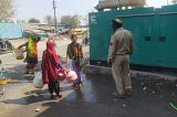 A photo with four people standing outside. Three are women wearing colourful clothing. They are walking away from the camera holding buckets in their hands. The fourth person is a security guard who is blocking them. They stand next to a grey road and a large green generator.