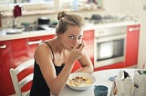 A blonde woman eats a bowl of oatmeal. She is wearing a black sleeveless camisole and sitting at a white kitchen table. The cabinets behind her are red.
