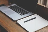 macbook and notepad with pen sitting on desk next to eachother