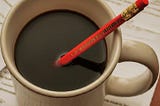A red pencil sits dunked in to a ceramic off-white mug full of black coffe