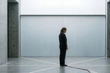 A single figure — gender neutral, mid-40s, sharp professional dress — standing in a vast empty space, architectural in feel. Concrete or glass. Six faint geometric lines or frequencies radiating outward from the figure — subtle, structural, not decorative. One of the lines is visibly interrupted — fractured mid-transmission. The figure is not in distress. They are still. The fracture is in the architecture around them, not in their expression. Lighting: cool, precise, slightly clinical. No text