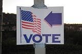 Photo displays a sign duct-taped to a pole with an American flag on one side and a left-facing arrow above the word “vote.”