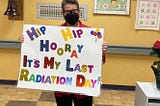 CC holding up a sign that reads, Hip Hip Hooray, It’s my last radiation day! Standing in front of the bell she is about to ring to celebrate the end of radiation!