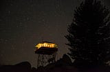 A fire lookout tower at night, with lights on inside, against a starry sky