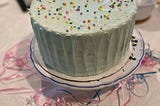 A mint green frosted cake with rainbow sprinkles on a cake stand with ribbons underneath
