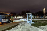 A blue and yellow SMART bus approaches a shelter at night with Oakland University’s clock tower in the background