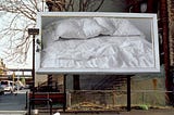 Photo of billboard with a picture of bed featuring two pillows with indentations where heads would have been.