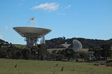 Deep Space Network antennas at Canberra Deep Space Communications Complex in Canberra Australia. The antenna in the foreground is pointed straight up at the sky. Two background antennas are pointed in different directions.