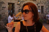 Photo of me, white female, mid-lengh dark-blond hair, on a black blouse and beaded necklace. Looking sideways with sunglasses, sniffing some wine on a glass. On the background, a stone wall from an old construction, two windows with dark wood frames. Some people seated on other tables behind me.