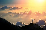 Silhoutte of man on top of a mountain against a background of sky filled with clouds and sun light rays.