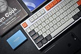 A white mechanical keyboard arranged side-by-side with a portable SSD, books and an iPhone.