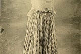 Black and white photograph from 1912 depicting a Moorish woman wearing a white blouse and flowy pants. Her arms are lifted. She is wearing bangles and a headdress composed of a light veil and jewelry.