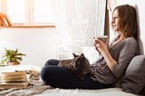 Young smiling woman sitting on a bed, sipping tea with a cat on her lap and a book by her feet | Neurodivergent Life