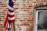 An American flag is tangled around a metal pole in front of a brick wall marked by white paint and a window with peeling frame.