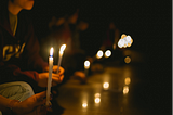 A hand holding a candle with a flame, with another person holding a lit candle also sitting in the background.